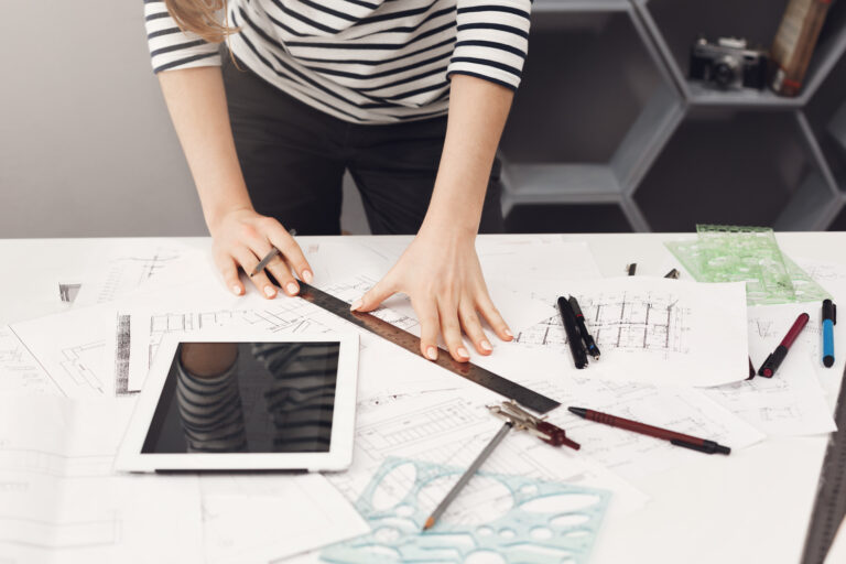 Top view of young good-looking architect student girl in casual striped shirt and black jeans standing near table, holding ruler and pen in hands making drawings, watching movie on digital table, getting ready for exams